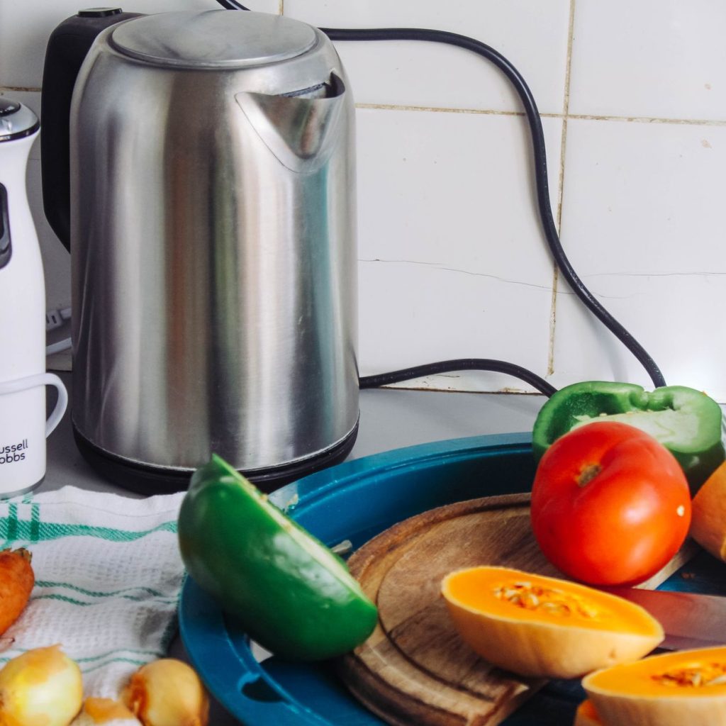 A vibrant array of fresh vegetables with a kettle and a blender on a kitchen countertop, ideal for healthy cooking. — Basic G