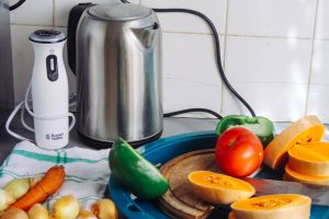 A vibrant array of fresh vegetables with a kettle and a blender on a kitchen countertop, ideal for healthy cooking. — Basic G