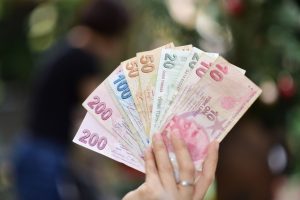 A hand holding various Turkish Lira banknotes with a blurred outdoor background. — Check Out The Superb Collection Of Vape Ju