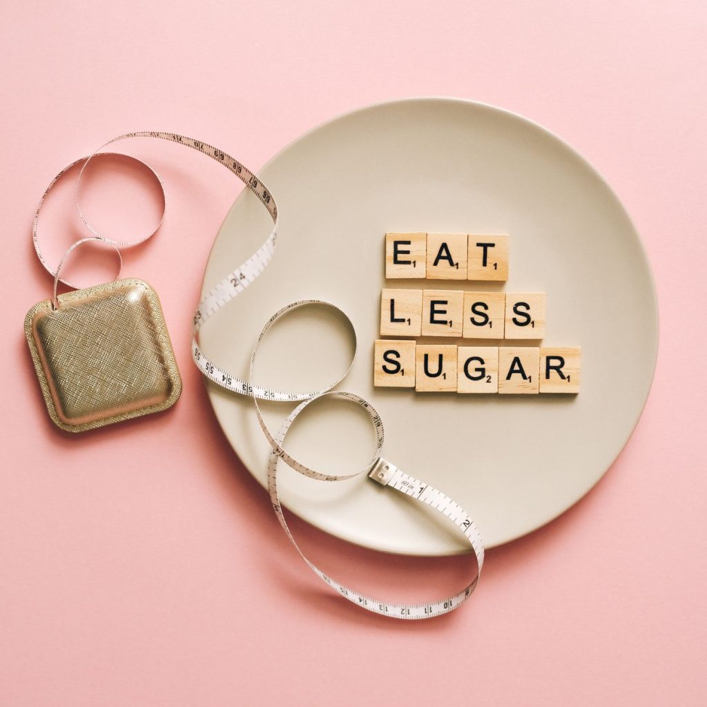 Flat lay of Scrabble tiles saying 'Eat Less Sugar' on a plate with a tape measure on a pink background. — Diets and Their Iss