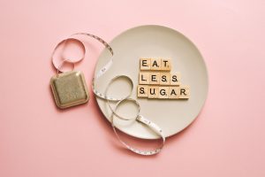 Flat lay of Scrabble tiles saying 'Eat Less Sugar' on a plate with a tape measure on a pink background. — Diets and Their Iss