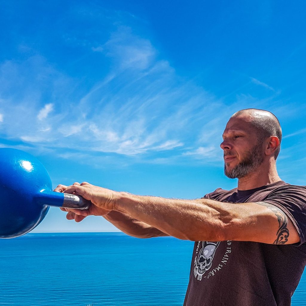 A fit, tattooed man lifting a kettlebell outdoors by the ocean under a clear blue sky. — Health and Fitness Exercises - Overv