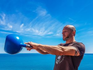 A fit, tattooed man lifting a kettlebell outdoors by the ocean under a clear blue sky. — Health and Fitness Exercises - Overv
