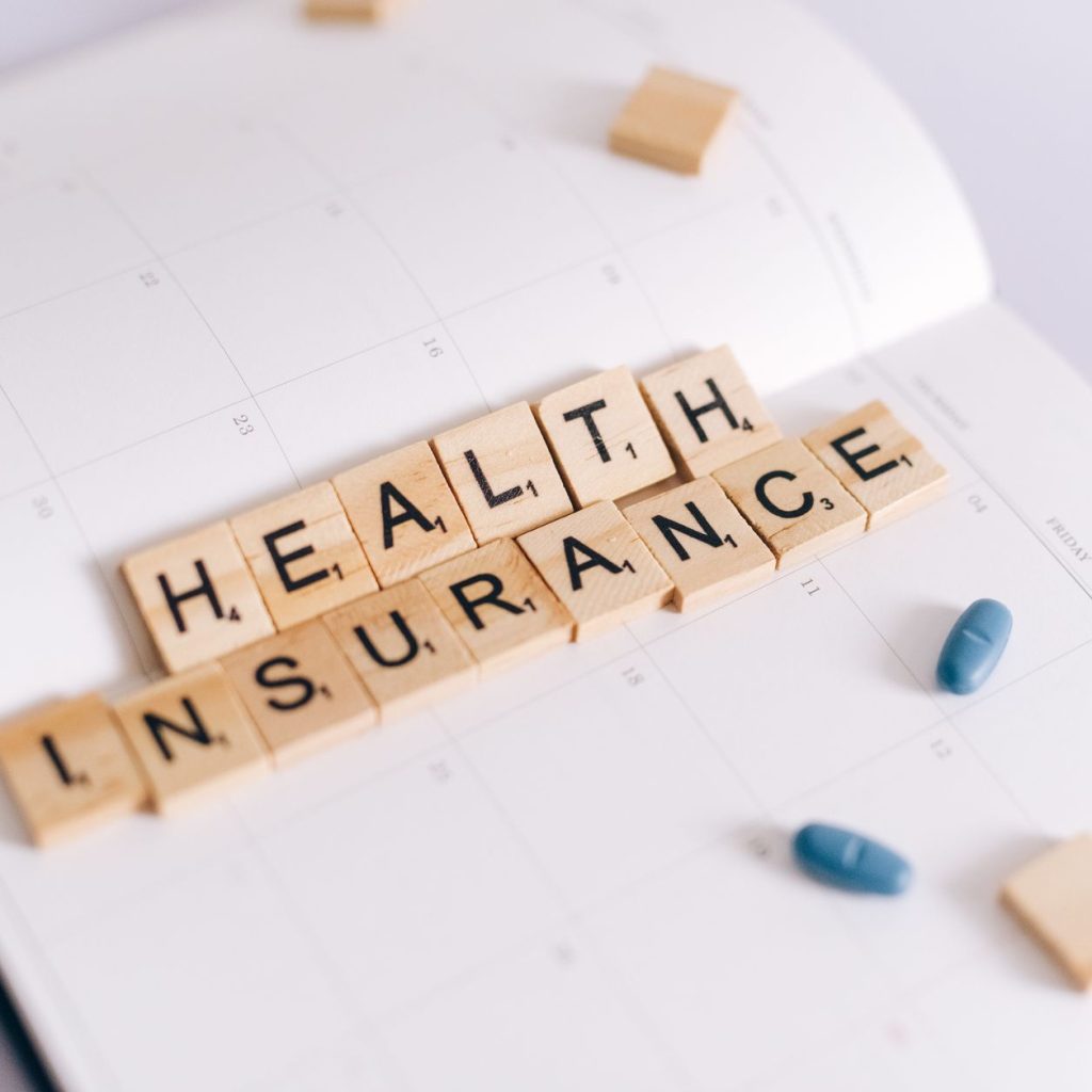 Close-up of health insurance spelled out with Scrabble tiles on a planner, alongside medication pills. — How To Spot On The M