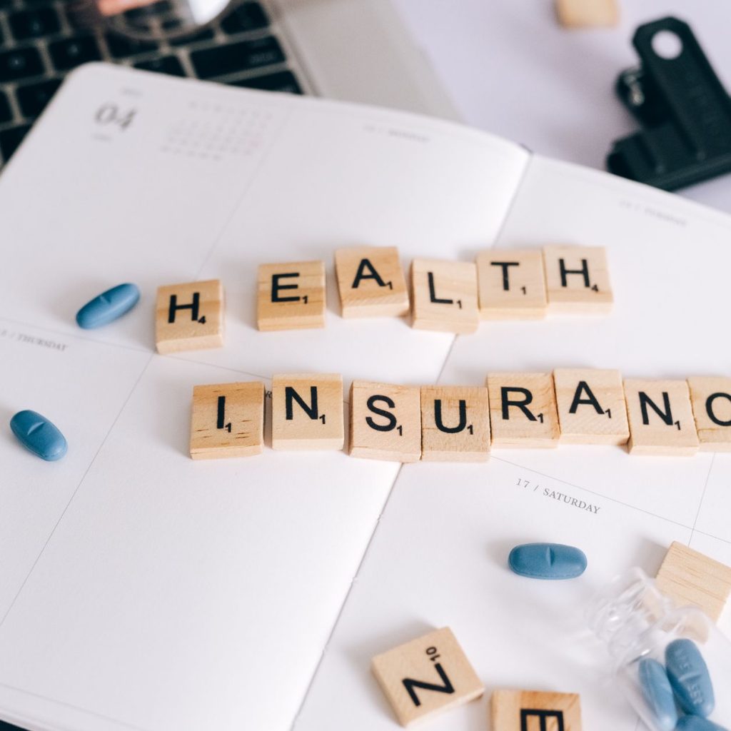 Scrabble tiles spelling 'Health Insurance' on planner with pills and laptop, symbolizing healthcare planning. — Seven Princip