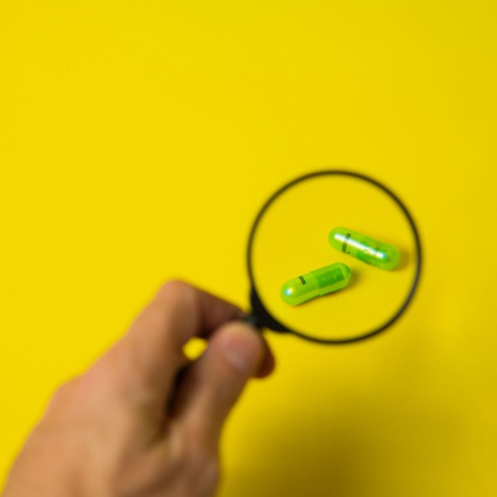From above of crop unrecognizable male holding magnifying glass over green pills on yellow background in studio — Why
