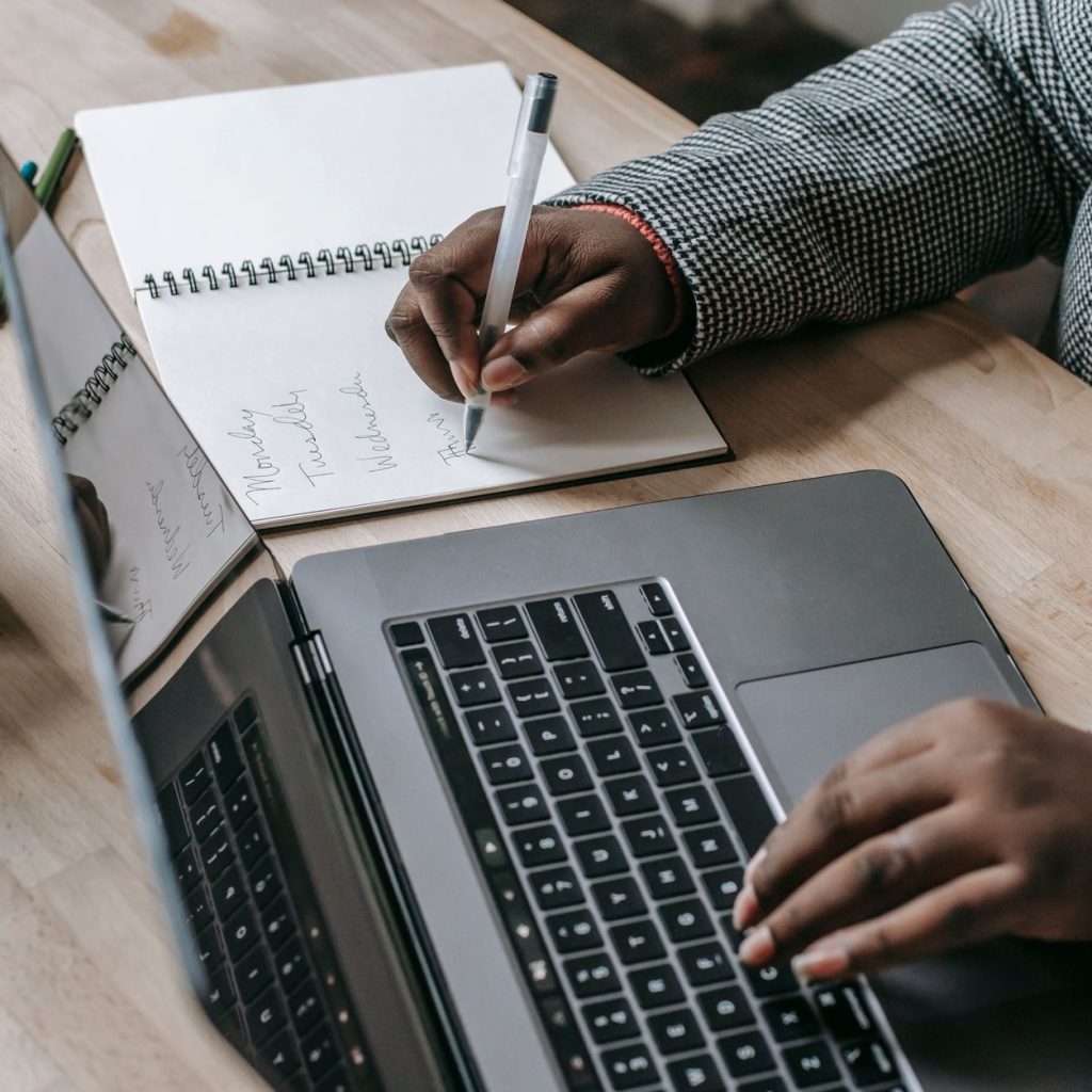 Crop unrecognizable African American female in formal wear writing notes in notepad while working on laptop — The Best Place