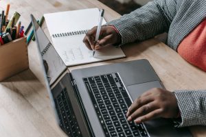 Crop unrecognizable African American female in formal wear writing notes in notepad while working on laptop — The Best Place