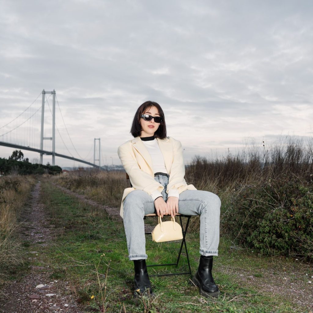 Stylish woman in sunglasses sitting by a bridge, showcasing modern fashion against a natural backdrop. — Things You Need to K