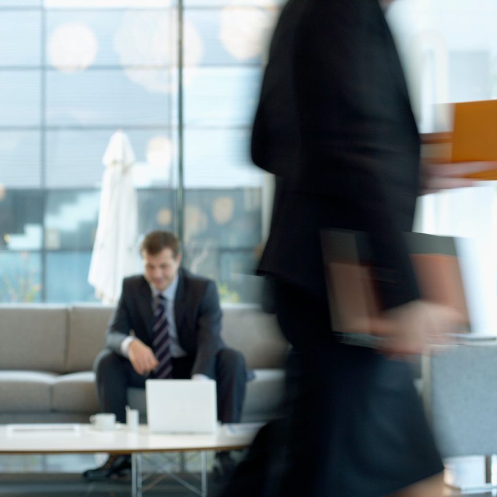 Businessman sitting in modern office lounge using a laptop while colleague walks past, creating dynamic blur. — What is an Ev