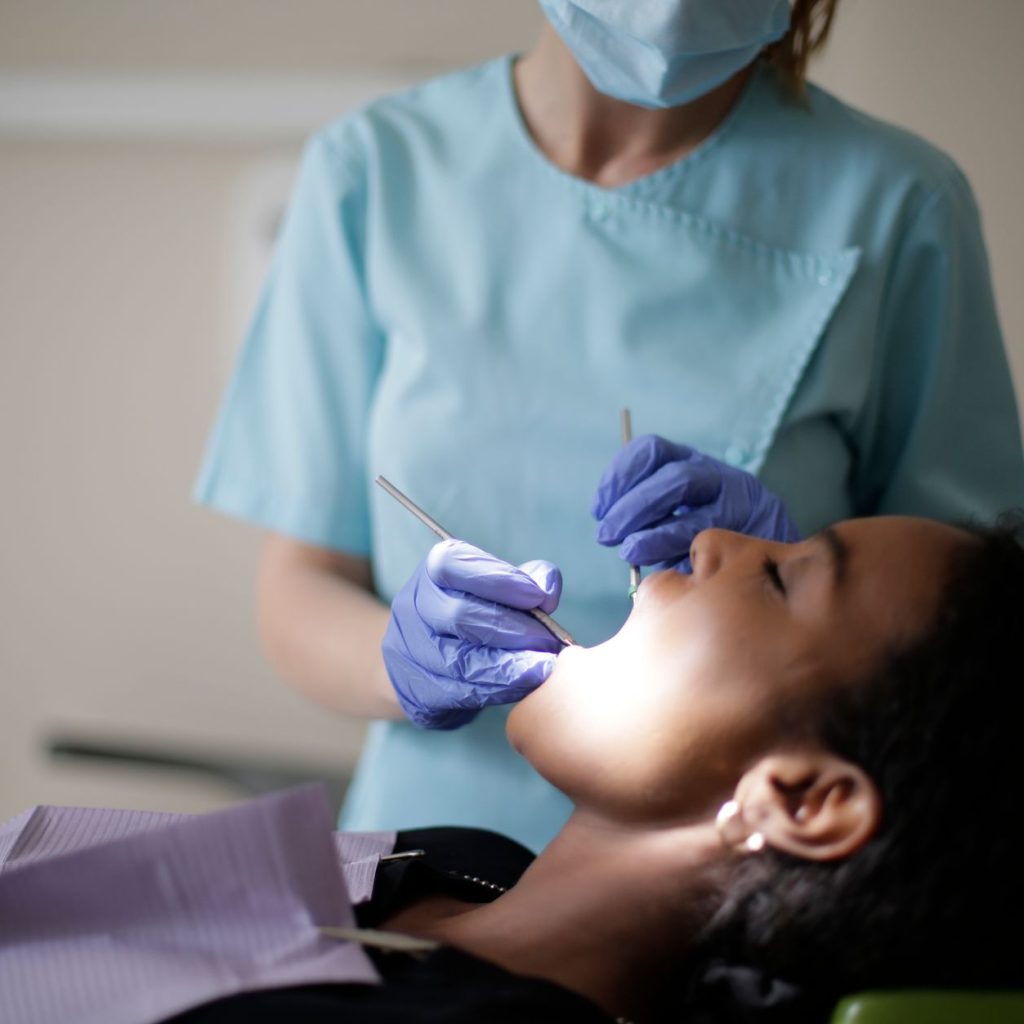 Focused female dentist in mask uniform and latex gloves using instruments for examining teeth of African American lady while