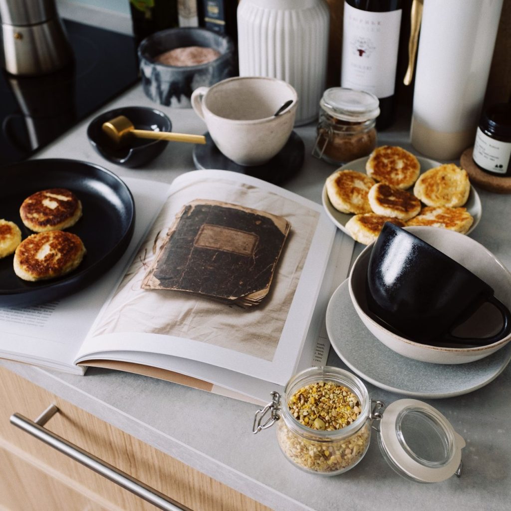 High angle of cookbook and cooked fritters on plates placed on table near utensils and ingredients in cozy kitchen at home —