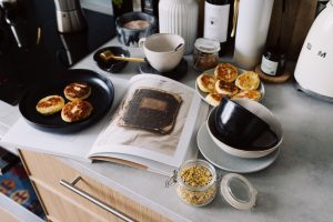 High angle of cookbook and cooked fritters on plates placed on table near utensils and ingredients in cozy kitchen at home —